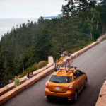 A car carrying bicycles drives on a narrow road overlooking the coastline.